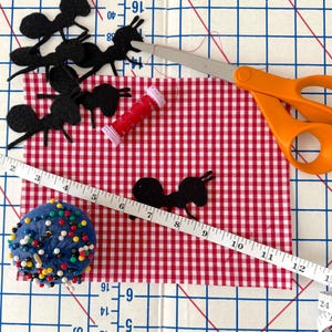 May include: A red and white gingham fabric rectangle featuring several black felt ant cutouts, a small spool of red thread, orange-handled scissors, a blue pincushion with colorful pins, and a white measuring tape showing up to 13 inches.