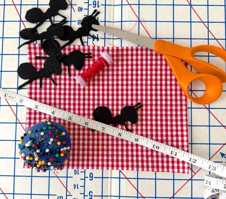 May include: A red and white gingham fabric rectangle featuring several black felt ant cutouts, a small spool of red thread, orange-handled scissors, a blue pincushion with colorful pins, and a white measuring tape showing up to 13 inches.