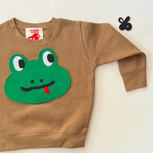 May include: A brown sweatshirt featuring a large green frog face applique on the front. The frog has black eyes, a black mouth, and a red tongue. The sweatshirt has a crew neck and long sleeves. A small tag with the text "Whimsy Ranch" is visible.