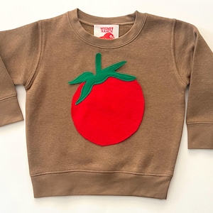 May include: A brown sweatshirt with a large red tomato applique and green leaves. The sweatshirt has long sleeves and a crew neck. The brand label "Wiggly Ranch" is visible on the neckline.