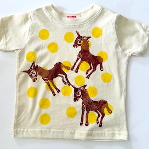 May include: A cream-colored short-sleeve t-shirt featuring a repeating pattern of bright yellow polka dots and three dark brown donkey illustrations. The inner neck label displays the text Whimsy in red lettering on a white background.