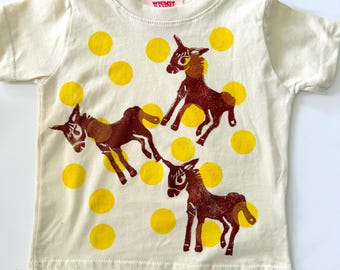 May include: A cream-colored short-sleeve t-shirt featuring a repeating pattern of bright yellow polka dots and three dark brown donkey illustrations. The inner neck label displays the text Whimsy in red lettering on a white background.
