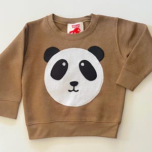 May include: A brown sweatshirt featuring a panda face applique. The panda has a white face with black eyes, ears, and a small nose. The sweatshirt has long sleeves and a crew neck. The tag reads "Whimsy Ranch".