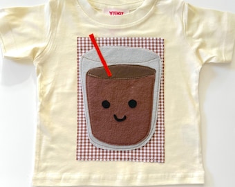 Puede incluir: Una camiseta de manga corta color crema con un parche rectangular de cuadros vichy marrones y blancos. En el centro, una aplicación de fieltro muestra un vaso con una bebida marrón, una pajita roja y una cara sonriente.