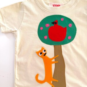 May include: A cream-colored short-sleeve t-shirt featuring a felt applique of a brown tree with a green canopy, a red bird, and an orange cat climbing the trunk. The inner neck label displays the text WHIMSY in red capital letters.