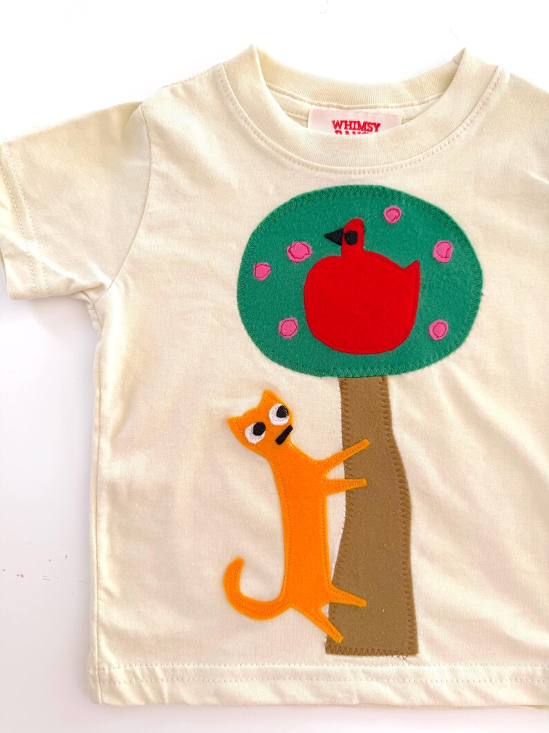 May include: A cream-colored short-sleeve t-shirt featuring a felt applique of a brown tree with a green canopy, a red bird, and an orange cat climbing the trunk. The inner neck label displays the text WHIMSY in red capital letters.