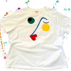 May include: White t-shirt with a colorful abstract face design. The design includes a green circle for an eye, a blue line for the nose, a yellow circle, and a red heart-shaped mouth. The shirt has short sleeves and a crew neck.