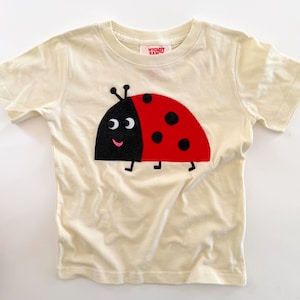 May include: A cream-colored short-sleeve t-shirt featuring a large ladybug graphic on the front. The ladybug has a black head, a red body with black spots, and a smiling face. The inner neck label displays the text Whimsy Ranch in red letters.