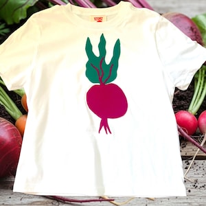 May include: A white t-shirt featuring a graphic of a beet with green leaves and a magenta root. The shirt is displayed on a wooden surface with fresh beets and radishes in the background. The shirt has a small tag on the collar.