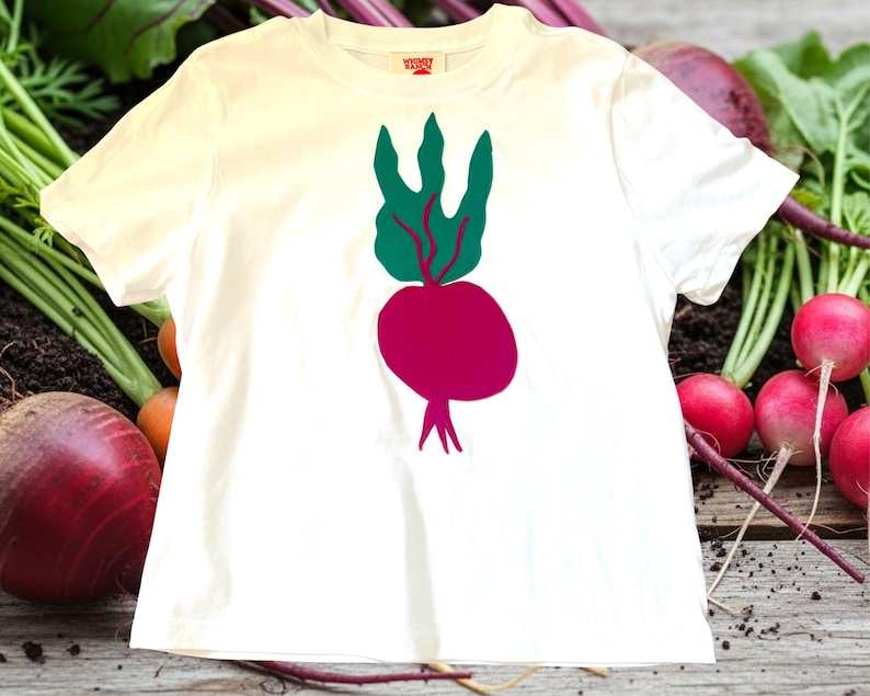 May include: A white t-shirt featuring a graphic of a beet with green leaves and a magenta root. The shirt is displayed on a wooden surface with fresh beets and radishes in the background. The shirt has a small tag on the collar.