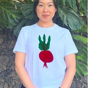 May include: White t-shirt featuring a red beet graphic with green leaves. The graphic is a simple, colorful illustration. The shirt is a classic crew neck style.