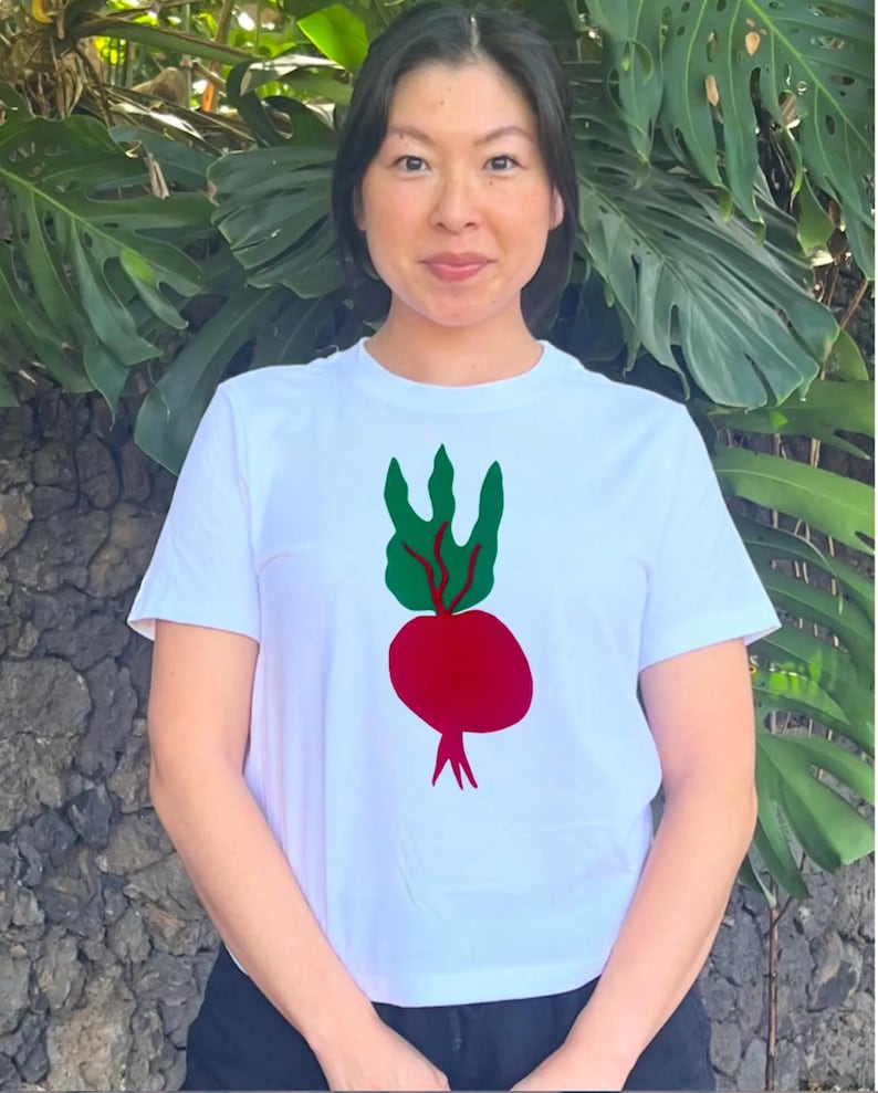 May include: White t-shirt featuring a red beet graphic with green leaves. The graphic is a simple, colorful illustration. The shirt is a classic crew neck style.
