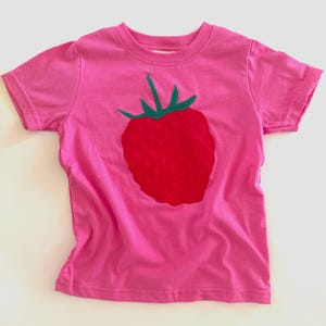 May include: A pink short-sleeved t-shirt featuring a large red strawberry applique with green leaves. The strawberry is centered on the front of the shirt. The shirt is made of a soft material.