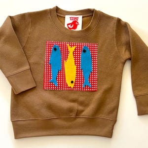 May include: Brown sweatshirt with a crew neck and long sleeves. The front features a red and white checkered patch with three felt fish: two blue and one yellow. The brand logo is a red bird.