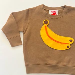 May include: A brown long-sleeve sweatshirt featuring a large yellow banana applique with orange stitching and brown accents. The interior neck label displays the text Whimsy Ranch with a red bird graphic. The garment has a crew neckline.