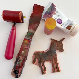 May include: A red rubber brayer, a metal palette knife with a dark handle, a small cylindrical yellow and red block, a tube of Speedball block printing ink, and a pink rubber stamp carved into the shape of a donkey on a white surface.