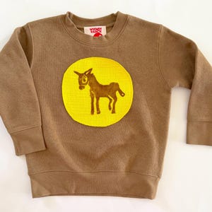 May include: A brown long-sleeve sweatshirt featuring a large, circular yellow patch on the chest. The patch displays a brown graphic of a donkey. The interior neck label features the text Whimsy Ranch in red lettering on a white background.