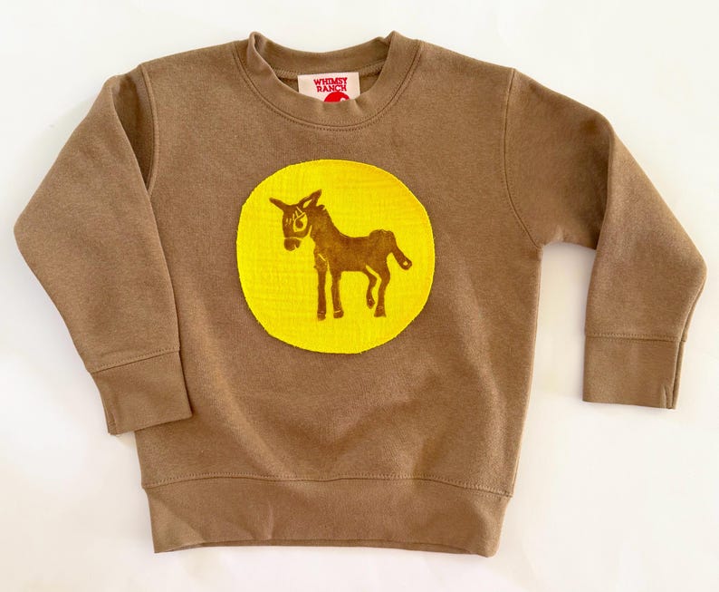 May include: A brown long-sleeve sweatshirt featuring a large, circular yellow patch on the chest. The patch displays a brown graphic of a donkey. The interior neck label features the text Whimsy Ranch in red lettering on a white background.