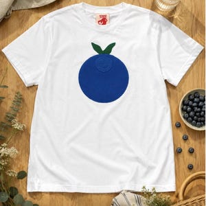May include: A white short-sleeve t-shirt featuring a large, solid blue circular graphic with two green leaves at the top. The inner neck label displays the text Whiskey Ranch in red. The shirt is constructed with a crew neckline and hemmed sleeves.