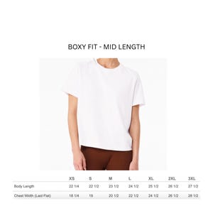 May include: A white, boxy fit, mid-length t-shirt. The image includes a size chart with body length and chest width measurements in inches for sizes XS to 3XL. The shirt is shown on a person wearing brown pants.