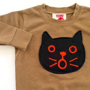May include: A long-sleeve brown crewneck shirt featuring a black cat face appliqu&eacute; with bright orange embroidered details. The shirt has a ribbed neckline and cuffs. A white label inside the collar displays the text WHIMSY RANCH in red.