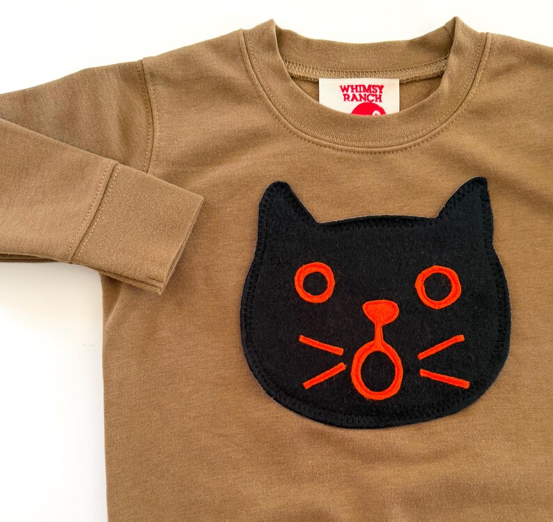 May include: A long-sleeve brown crewneck shirt featuring a black cat face appliqu&eacute; with bright orange embroidered details. The shirt has a ribbed neckline and cuffs. A white label inside the collar displays the text WHIMSY RANCH in red.