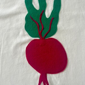 May include: A felt applique of a beet on a white background. The beet is a deep magenta color with green leaves and red stems. The leaves are a bright green color. The applique is likely sewn onto a piece of fabric.