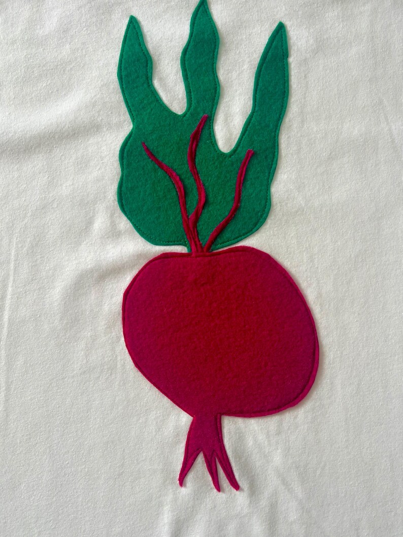 May include: A felt applique of a beet on a white background. The beet is a deep magenta color with green leaves and red stems. The leaves are a bright green color. The applique is likely sewn onto a piece of fabric.