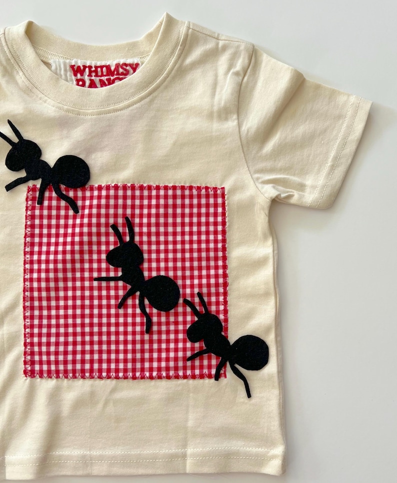 May include: A cream-colored short-sleeve t-shirt featuring a red and white gingham square patch stitched onto the front. Three black ant-shaped fabric appliqu&eacute;s are positioned across the patch and shirt. The inner neck label reads WHIMSY.
