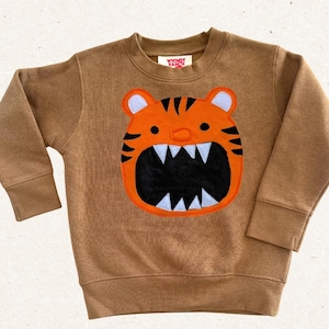 May include: A tan long-sleeve sweatshirt featuring a large orange tiger face appliqué on the front. The tiger has black stripes, white ears, and a wide-open mouth with white teeth against a black background. The inner neck label reads Whimsy Ranch.