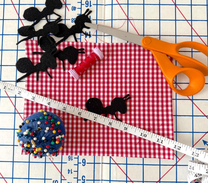 May include: A craft scene with black felt ant cutouts, red and white gingham fabric, orange scissors, a red spool of thread, a blue pin cushion, and a measuring tape. The measuring tape shows inches.