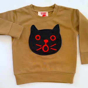 May include: A tan long-sleeve sweatshirt featuring a black cat face appliqu&eacute; with bright red embroidered eyes, nose, mouth, and whiskers. The interior neck label displays the text Whimsy Ranch in red letters on a white background.