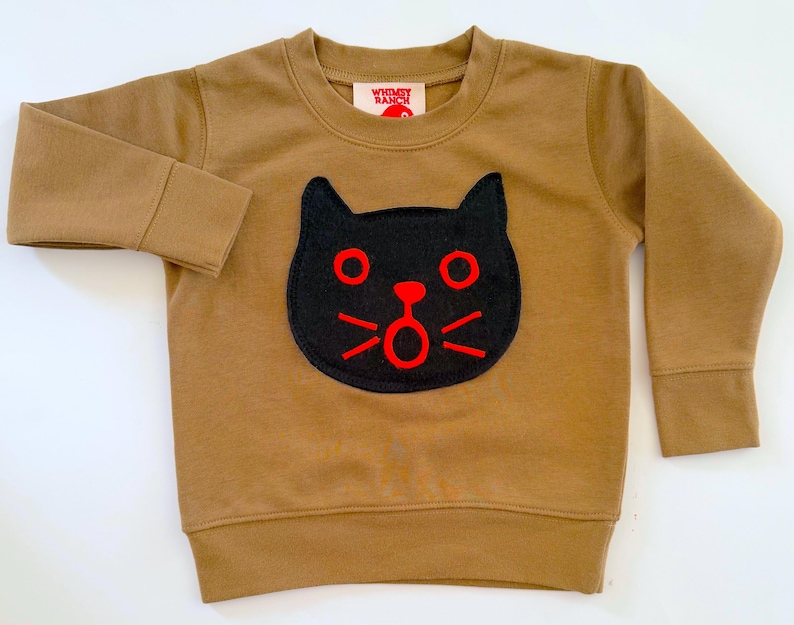 May include: A tan long-sleeve sweatshirt featuring a black cat face appliqu&eacute; with bright red embroidered eyes, nose, mouth, and whiskers. The interior neck label displays the text Whimsy Ranch in red letters on a white background.