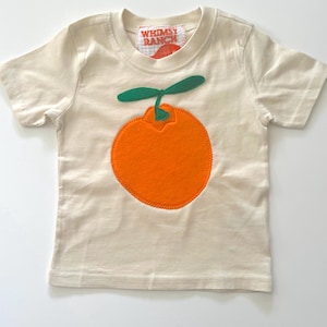 May include: A cream-colored short-sleeve t-shirt featuring a large, bright orange circular applique with a green leaf stem on the chest. The interior neck label displays the text Whimsy Ranch in orange on a white and orange checkered background.