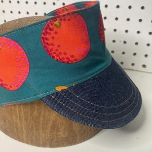 May include: A visor with a denim peak and a teal fabric band featuring large orange fruit designs with pink accents. The visor is set on a wooden form, showcasing its shape and construction.