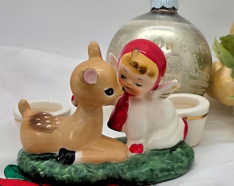 May include: A ceramic figurine featuring a fawn and an angel with a red hood and scarf. The angel is embracing the fawn, both resting on a green base. A silver Christmas ornament is in the background. The figurine is a Christmas decoration.