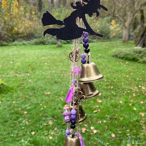 Könnte beinhalten: Ein Windspiel mit einer Silhouette einer Hexe mit lila und schwarzen Perlen, einem Schlüsselanhänger und vier goldenen Glocken. Das Windspiel hängt an einer braunen Schnur.