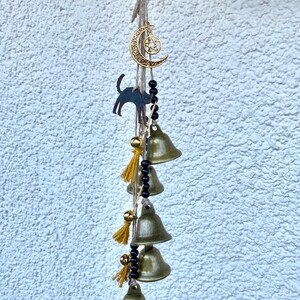 Pode incluir: Um amuleto de lua crescente e pentagrama dourado pendurado em uma corda de contas pretas e sinos dourados. O amuleto está preso a uma corda de juta marrom com borlas amarelas.