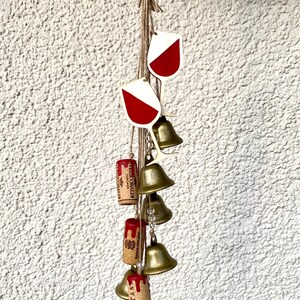 Op de afbeelding: Een windgong met wijnthema met rode en witte wijnglasbedels, gouden bellen en kurkaccenten. De kurken hebben wijnlabels met tekst zoals "Fontana" en "Frescobaldi Toscana".