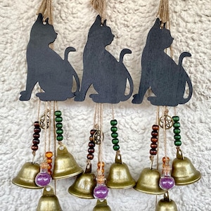 Pode incluir: Três sinos de vento com silhueta de gato preto, com contas marrons e verdes, sinos dourados e pequenos frascos de vidro com purpurina roxa. Cada sino de vento tem um charme de chave na parte inferior.