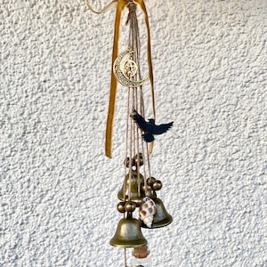 Pode incluir: Um móbile dourado e castanho com um amuleto de lua crescente, um amuleto de pássaro preto e uma pequena garrafa de vidro cheia de conchas. O móbile está pendurado em uma fita bege e um cordão castanho.