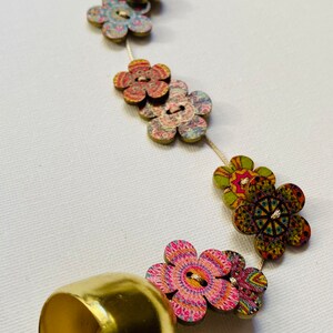 Pode incluir: Um sino dourado com uma corda de botões de flores de madeira coloridos. O sino está pendurado em um anel dourado.