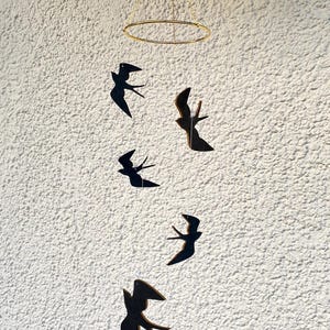 Peut inclure: Un anneau en métal doré avec une chaîne qui en pend. Cinq silhouettes d'oiseaux noires sont suspendues à la chaîne. Les silhouettes d'oiseaux sont dans différentes positions, certaines face avant et d'autres face arrière. L'arrière-plan est un mur blanc.