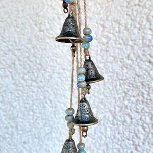 Könnte beinhalten: Eine Schnur mit sechs kleinen, antik aussehenden Glocken mit filigranen Designs hängt an einem Stück Schnur. Die Glocken sind mit blauen und weißen Keramikperlen durchsetzt.
