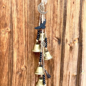 Pode incluir: Um móbile de vento dourado com um amuleto de lua crescente e pentagrama, um amuleto de corvo preto e cinco sinos dourados pendurados em um cordão de juta marrom com contas pretas.