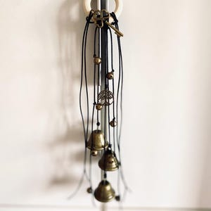 Könnte beinhalten: Ein dekorativer Windspiel mit einem Holzring, schwarzen Schnüren und bronzefarbenen Anhängern. Zu den Anhängern gehören ein Schlüssel, Glocken, Perlen und ein Lebensbaum-Anhänger. Das Windspiel hängt vor einem weißen Hintergrund.