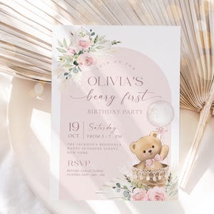 Pink floral first birthday invitation featuring a teddy bear in a basket, pastel flowers, and a balloon for a baby girl's Beary First birthday party