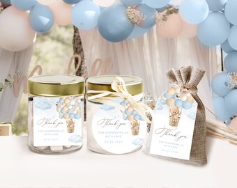 Może przedstawiać: Upominki na baby shower z misiem w balonie na gorące powietrze. Upominki obejmują szklane słoiki ze złotymi pokrywkami i jutową torbę, wszystkie z pasującymi etykietami.