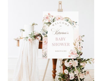 May include: A welcome sign for a baby shower featuring a floral arch design. The sign is white with pink and gold accents and reads "Welcome to Katarina's Baby Shower, March 10, 2023".