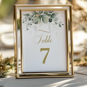 Op de afbeelding: Een ingelijst tafelnummer met een gouden lijst. De witte kaart heeft het woord "Table" in sierlijk schrift en het nummer "7" in een vetgedrukte letter. De bovenkant van de kaart is versierd met groene en gouden aquarelbladeren.